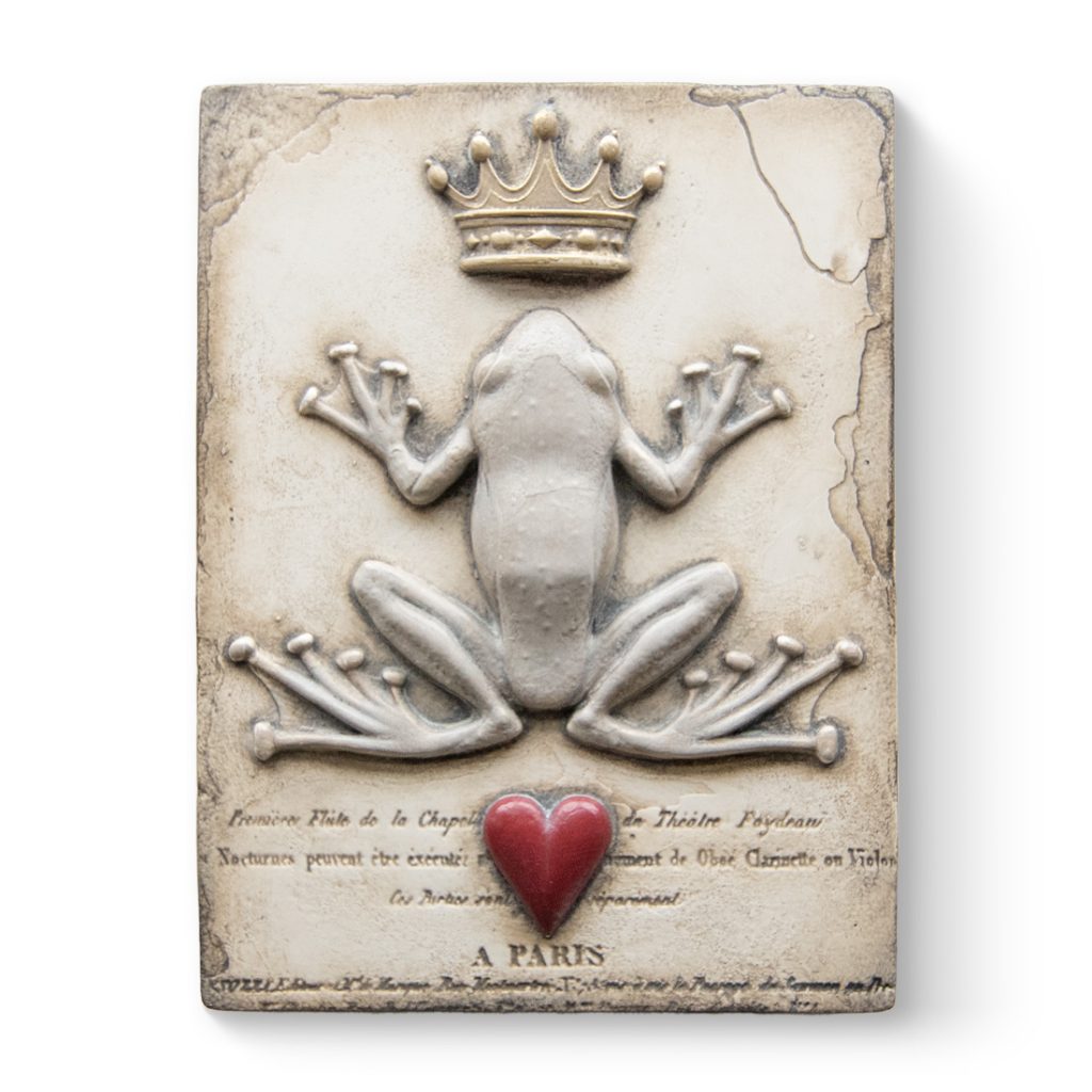 A white sculptural block a 3D crown, frog, and heart on it. The crown, frog, and heart are laid out in that order from the top of the block to the bottom. At the bottom of the block script text is carved into it underneath the heart.
