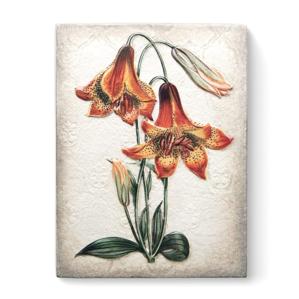 A white sculptural block with a vintage illustration of orange trumpet shaped flowers with long green stems and green leaves on it. The flowers start yellow on the inside becoming deep orange on the outside. Inside the flowers are black freckles and long stamen. The back of the block has a vintage wallpaper pattern craved into it.